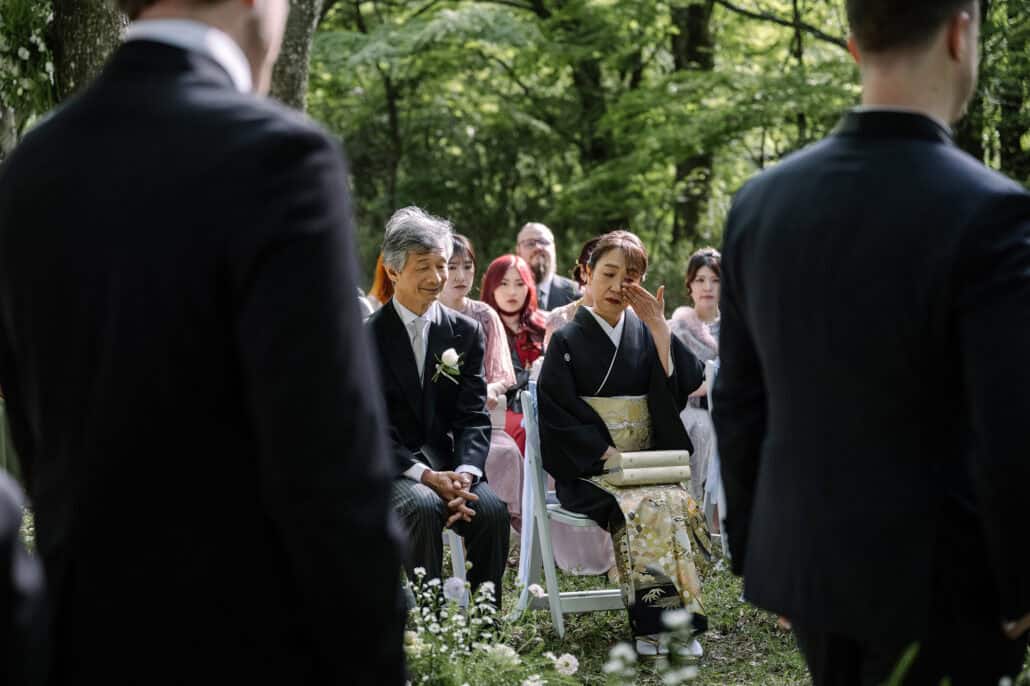 Her father in a morning coat smiles and her mother in a black kimono with a gold obi wipes an eye in the front row of the forest wedding ceremony.