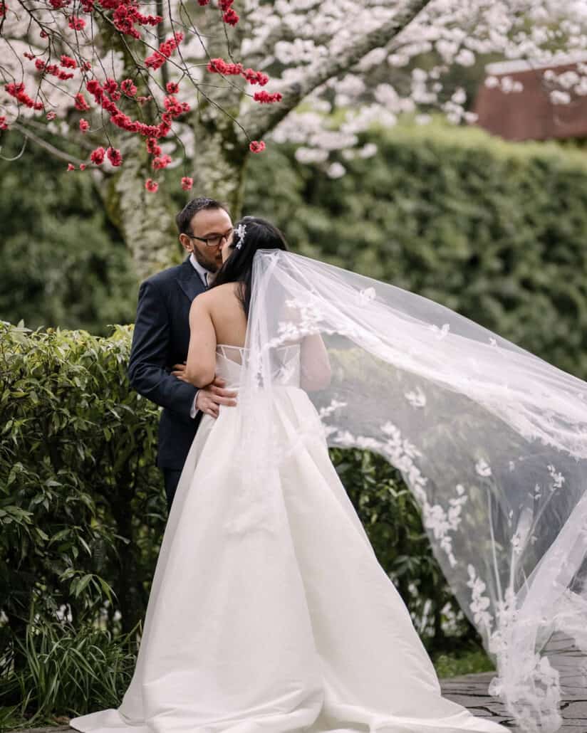 The bride and groom kiss beneath white cherry blossom and red spring flowers in Shuzenji, her long veil lifting on the wind.