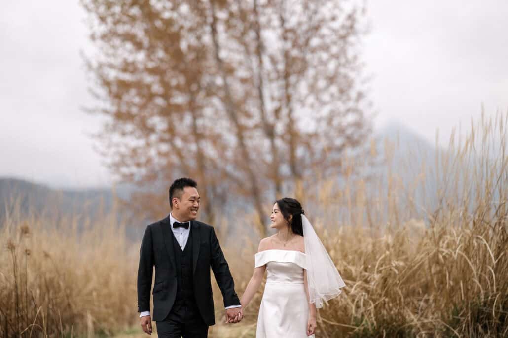 The bride and groom walk hand in hand through tall golden grass, smiling at each other, a tall brown tree and misty hills behind them.