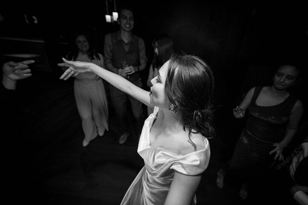 In black and white, the bride dances with one arm outstretched as smiling guests gather around her at the wedding reception at BANK30 in Takeshiba.