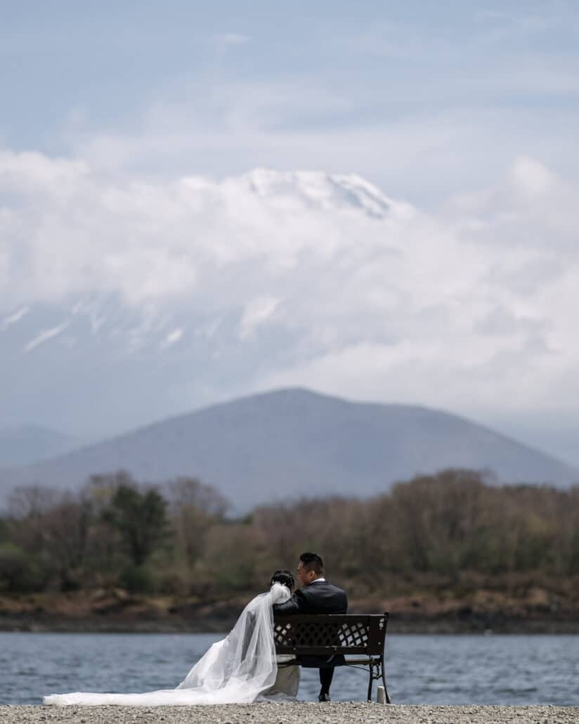 The bride and groom sit on a bench at the water's edge, her veil and train across the gravel, Mt Fuji's summit showing through the cloud above.
