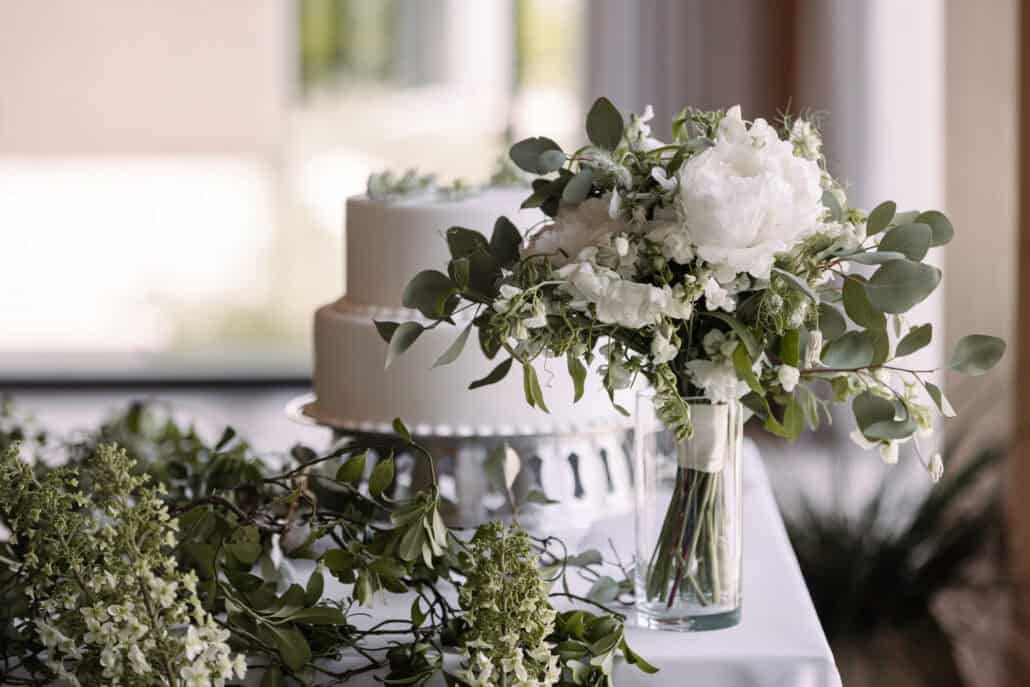 A two-tier white wedding cake on a silver stand beside the bride's bouquet of white peonies and eucalyptus in a glass vase, at Rosewood Miyakojima.