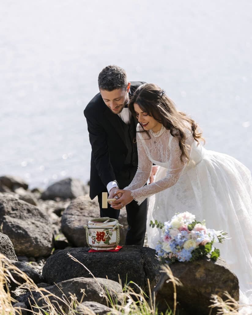 The bride and groom hold one wooden mallet together over a small sake barrel on dark lava rocks for kagami biraki on the shore of Lake Kawaguchi.