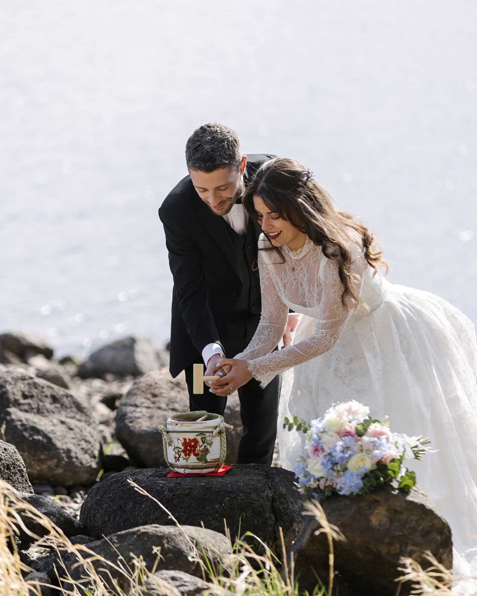 A bride and groom hold one wooden mallet over a small sake barrel on dark lava rocks for kagami biraki on the shore of Lake Kawaguchi.