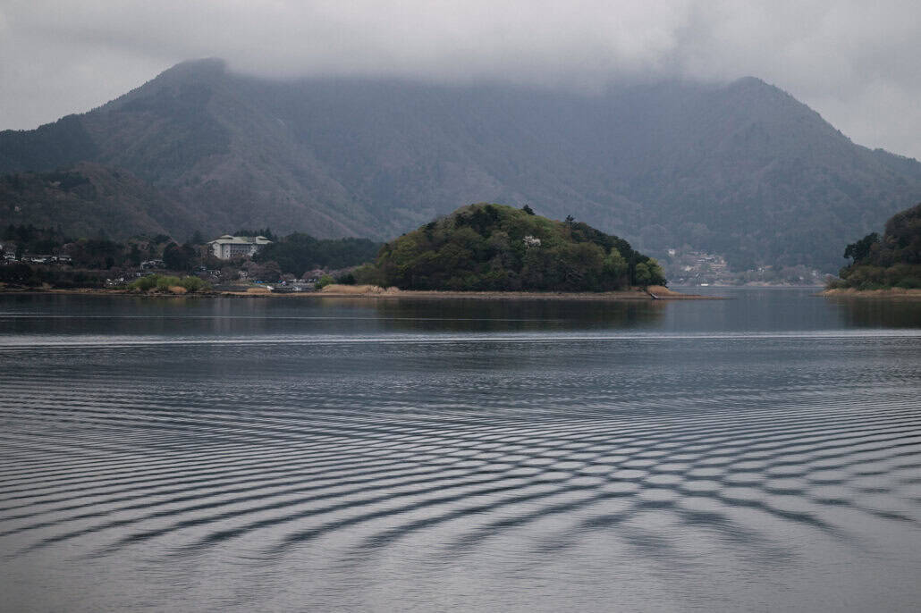 A small wooded island sits in Lake Kawaguchi below mountains wrapped in low cloud, crossed ripples patterning the grey water in front.