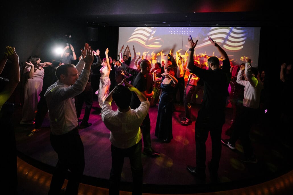 A packed dance floor with arms raised in coloured light before the projection wall, the bride among her guests at a wedding dance party in Tokyo.