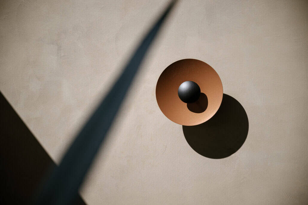 A round copper-toned wall light with a black sphere at its centre throws a circular shadow on warm plaster at Rosewood Miyakojima.