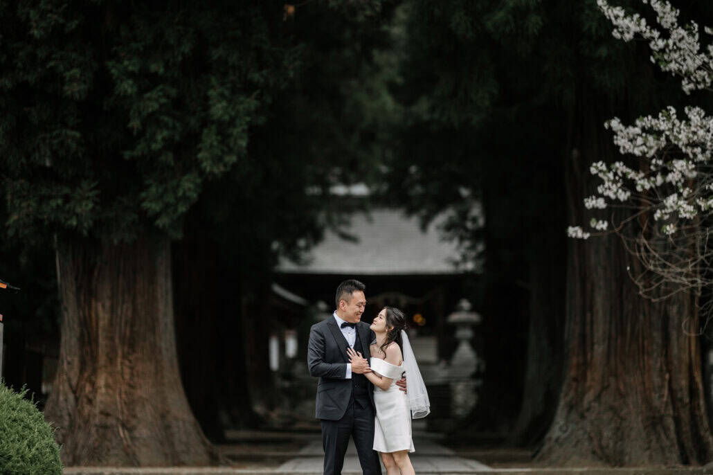 The bride and groom hold each other between two giant cedar trunks on the approach to Kawaguchi Asama Shrine, white blossom at the edge.