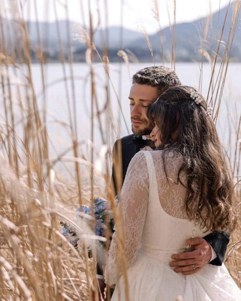 The groom closes his eyes as the bride leans into him among tall dry grass on the shore of Lake Kawaguchi, a pale blue bouquet low beside them.