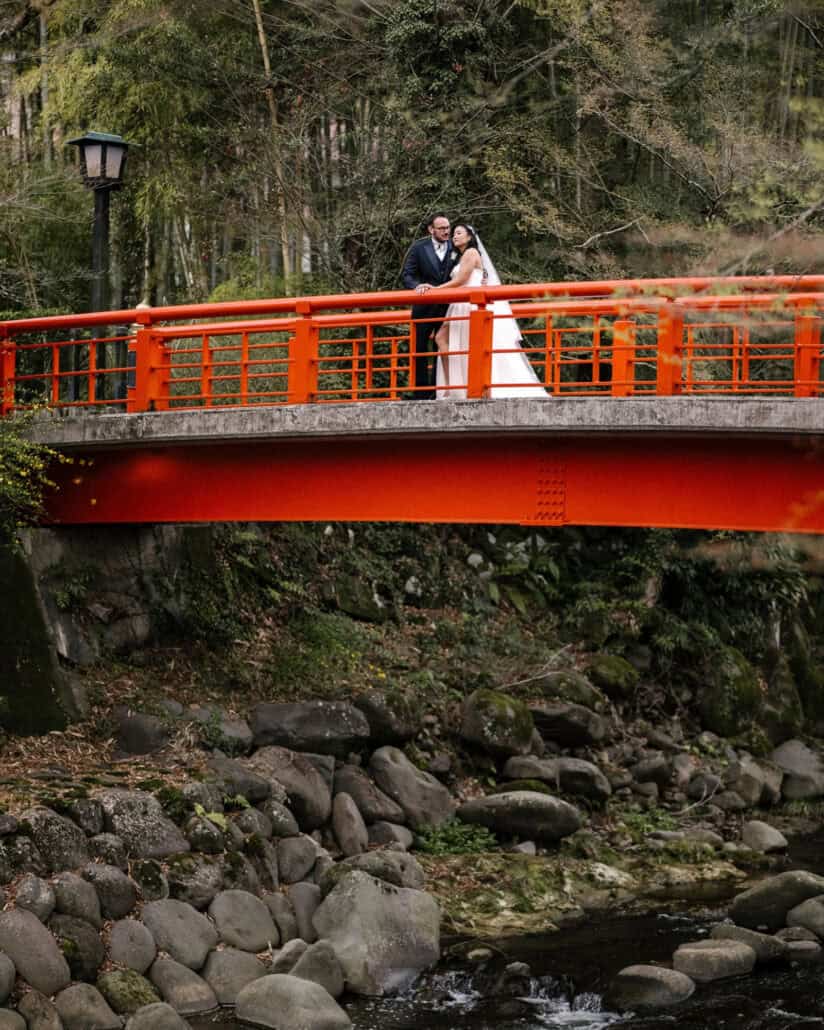 The bride and groom stand close at the rail of Shuzenji's red bridge for wedding portraits, a rocky river below and bamboo behind.