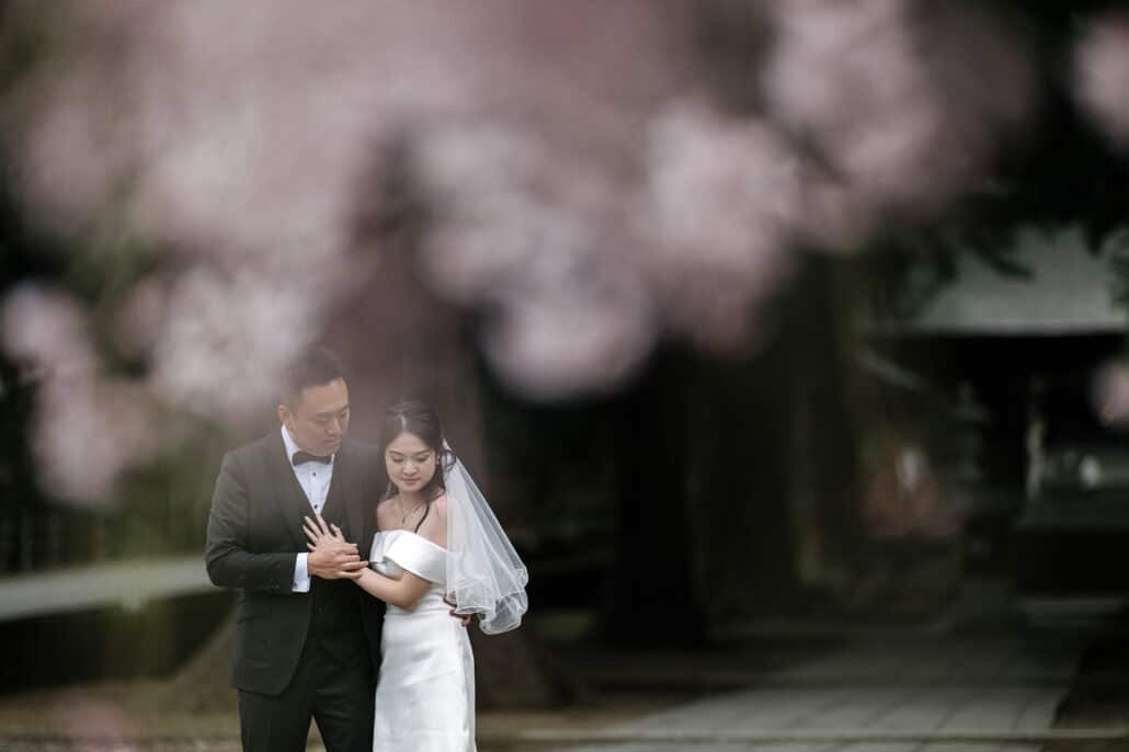 Pink cherry blossom blurs across the frame as the bride and groom hold each other on the stone approach to Kawaguchi Asama Shrine.