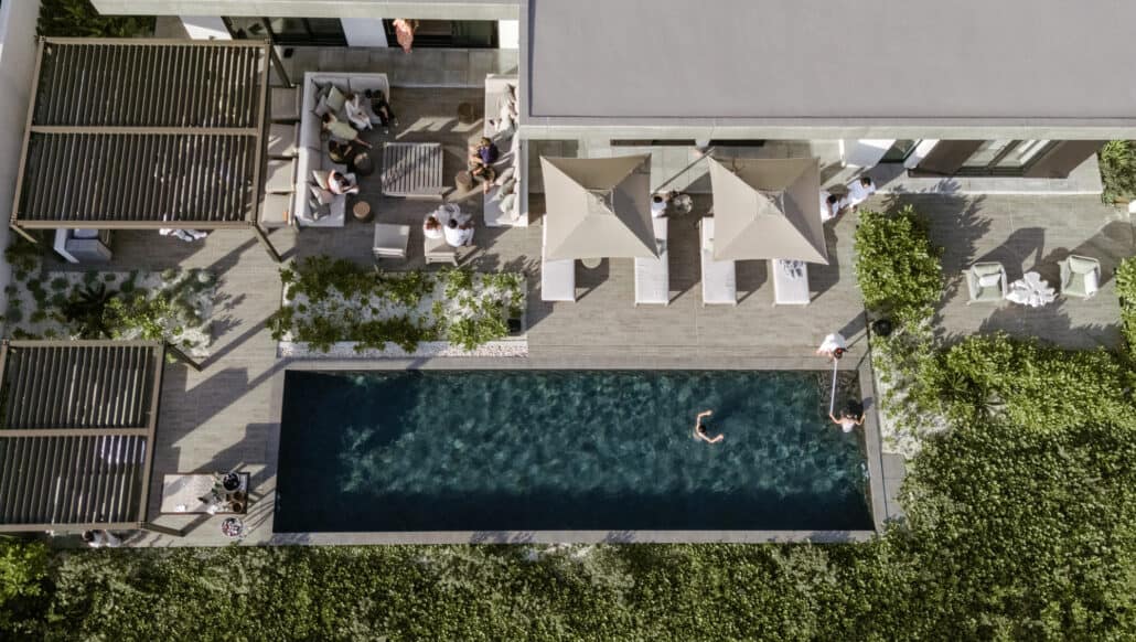 From above, wedding guests gather on sofas and under cream parasols beside a long dark pool on a terrace at Rosewood Miyakojima, one of them swimming.