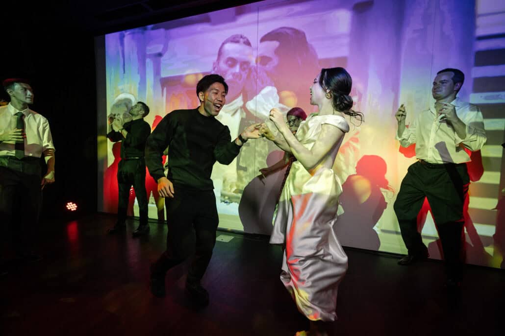 The bride dances hand in hand with a laughing guest in front of the projection wall at her wedding reception in Tokyo, friends dancing either side.