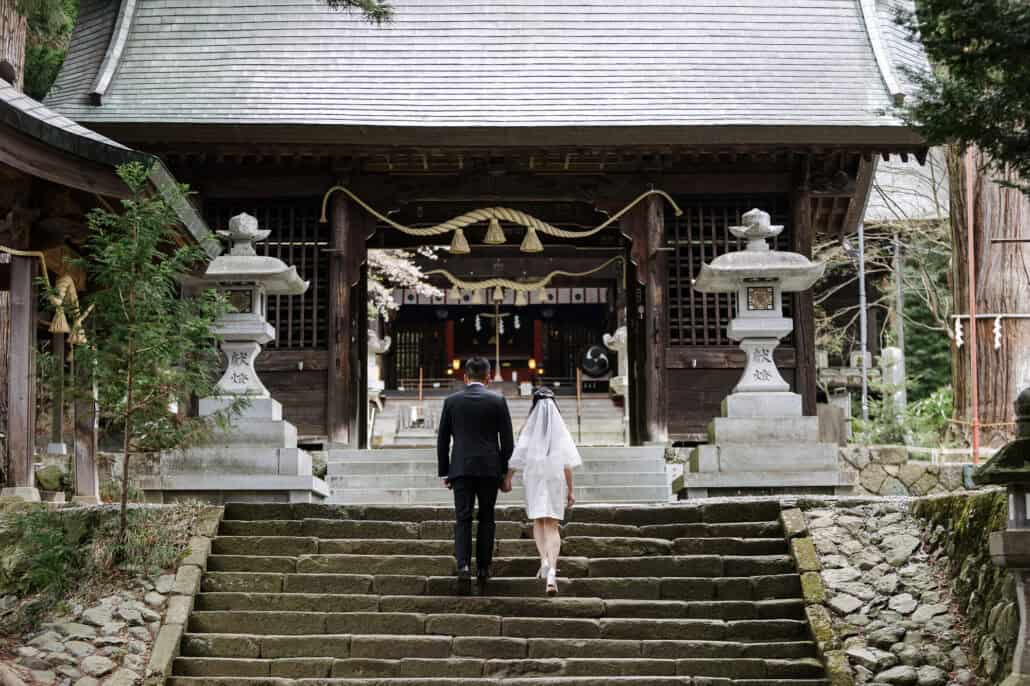 The bride and groom climb worn stone steps hand in hand to the gate hall of Kawaguchi Asama Shrine, a stone lantern on either side.
