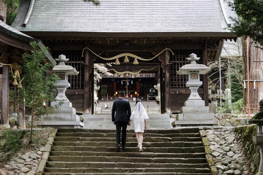 The bride and groom climb worn stone steps hand in hand to the gate hall of Kawaguchi Asama Shrine, a stone lantern on either side.