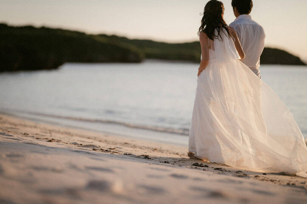 The bride and groom walk along the water's edge in low evening light, her sheer skirt lifting in the wind, toward a wooded headland on Miyako Island.