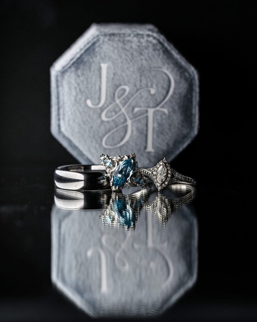 The wedding rings, one set with blue stones, sit reflected on a glossy black surface in front of a grey velvet box engraved with initials.