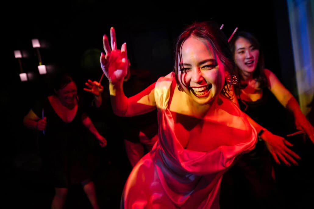 The bride laughs straight at the camera in red light with a glow stick in her hair as friends dance behind her at the wedding after party in Tokyo.
