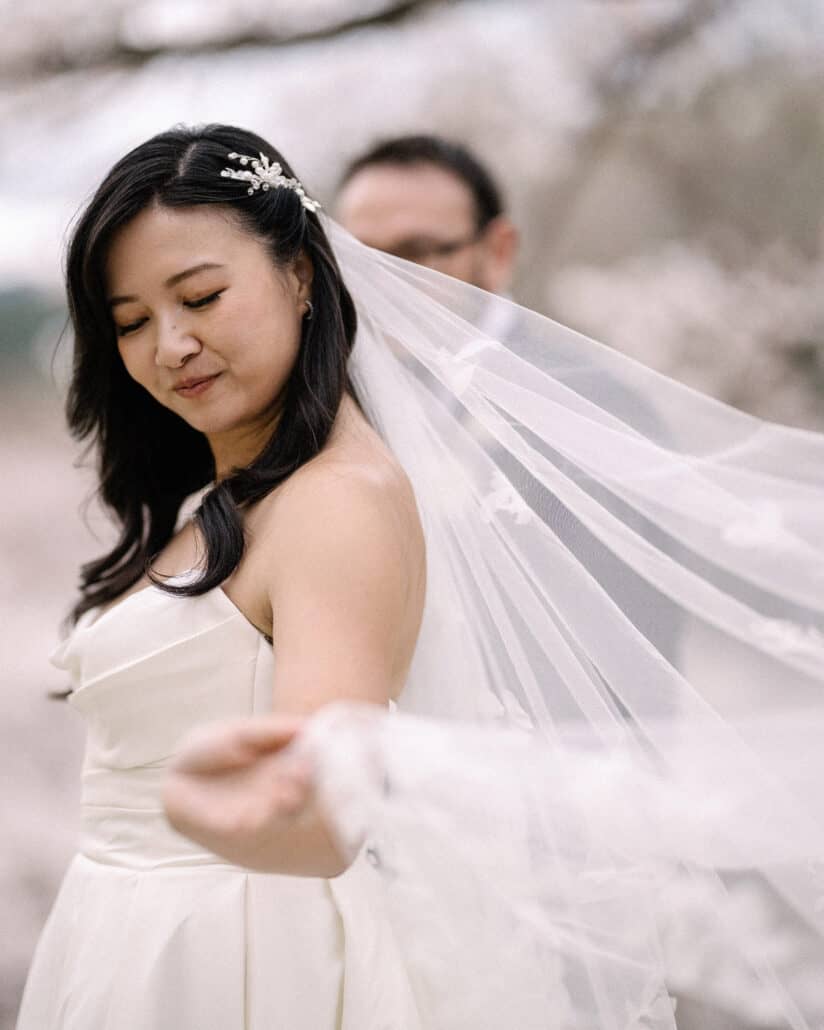 The bride smiles and draws her sheer veil out with one hand, the groom soft in focus behind her, under cherry blossom near Shuzenji.