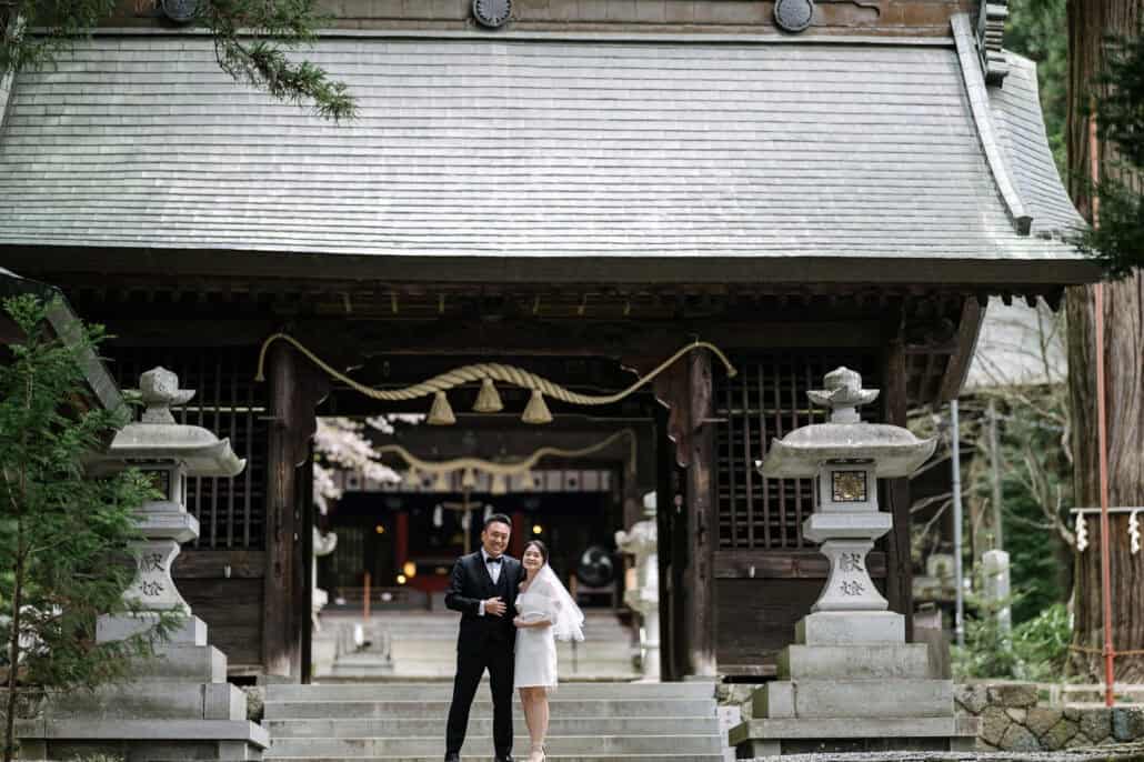 The bride and groom stand smiling below the gate hall's thick straw rope at Kawaguchi Asama Shrine, a stone lantern on either side of the steps.