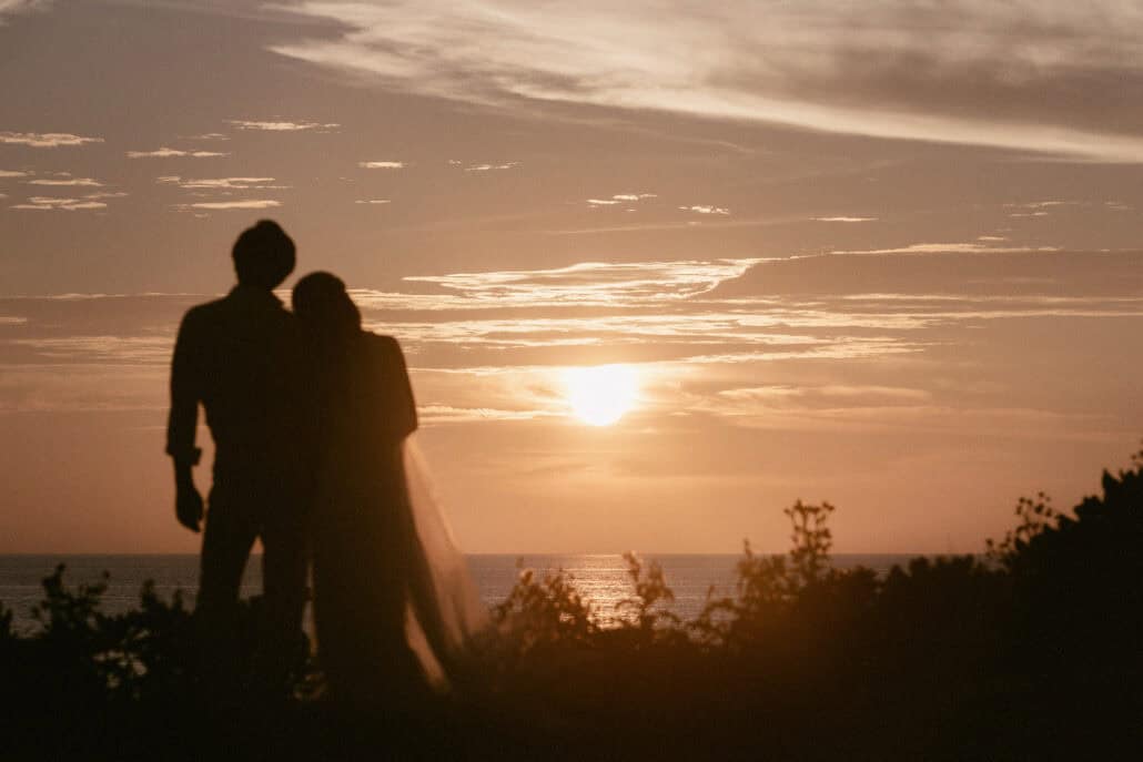 The couple stand close in silhouette on a clifftop, her veil hanging behind her, as the sun sinks through cloud at an Okinawa destination wedding.