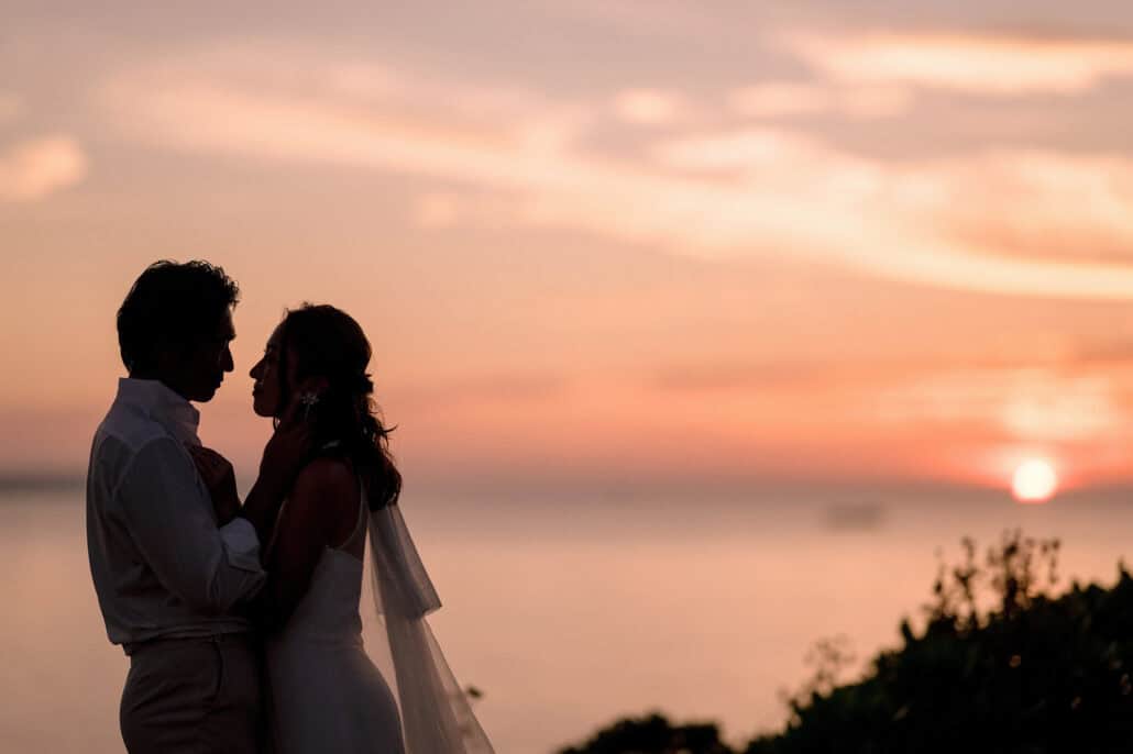 The bride and groom stand face to face in silhouette, her hand at his cheek, as the sun sits just above the sea at their Miyakojima wedding.