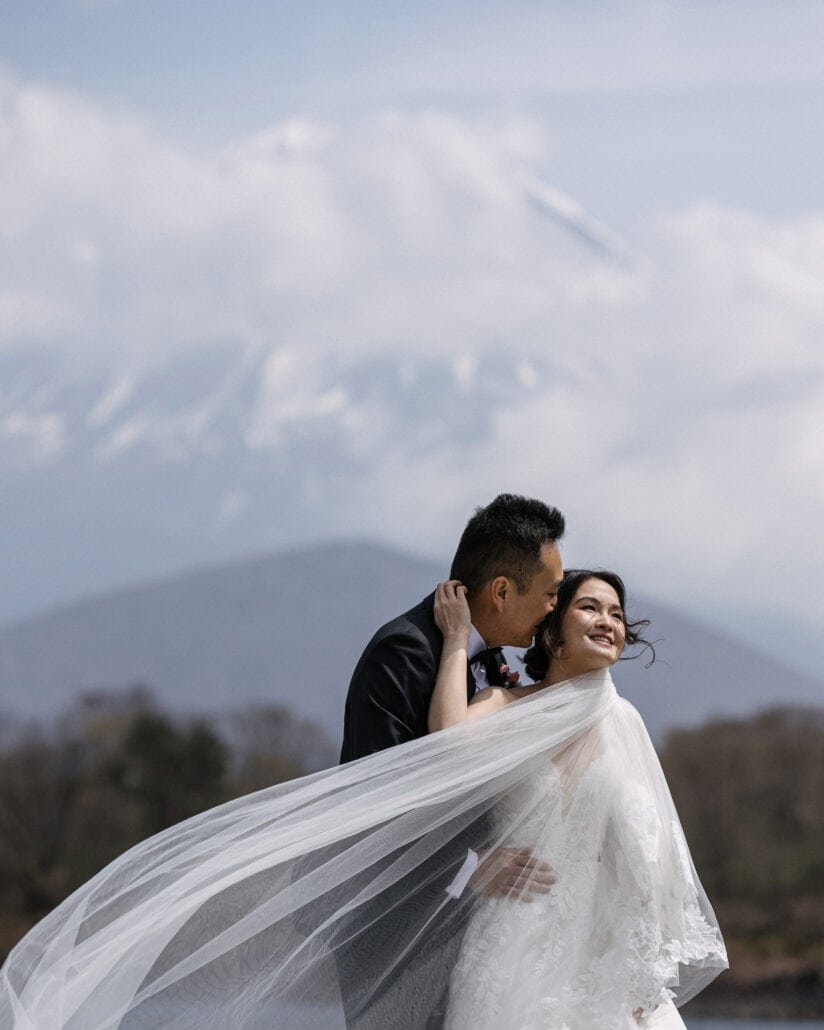 The groom kisses the bride's temple as her veil streams in the wind by Lake Shoji, the snow of Mt Fuji showing through the cloud above.