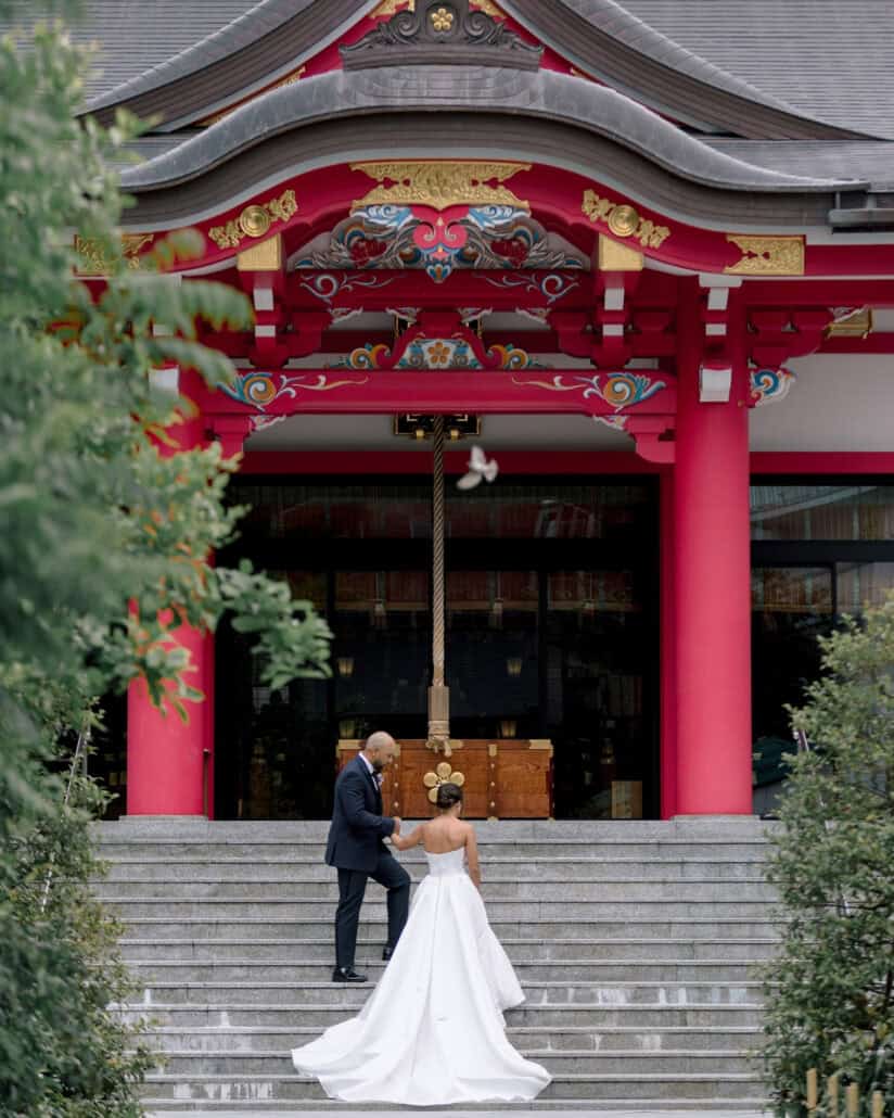 The groom helps the bride up the stone steps of a vermilion shrine hall for their Shinto wedding blessing in Tokyo.