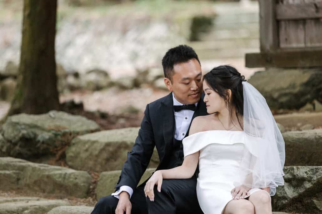 The bride and groom sit close on rough stone steps in the grounds of Kawaguchi Asama Shrine, heads together and eyes lowered.