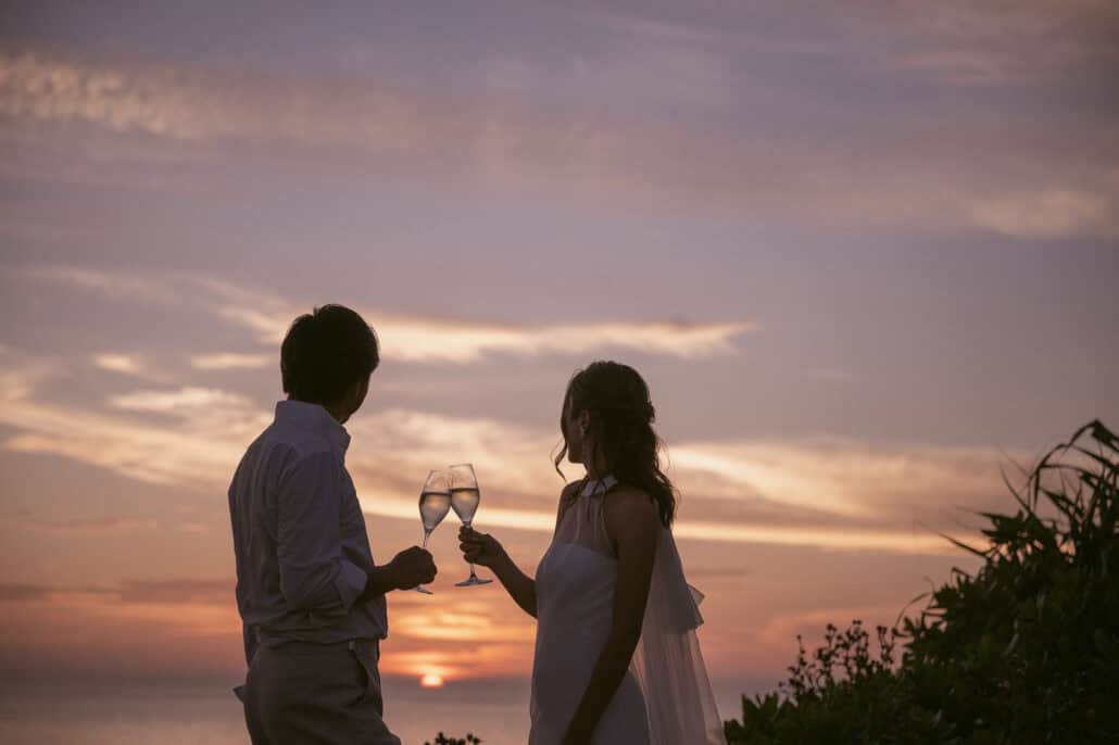 The bride and groom touch champagne glasses on a clifftop as the sun meets the sea under a pink and violet sky, at their destination wedding in Japan.