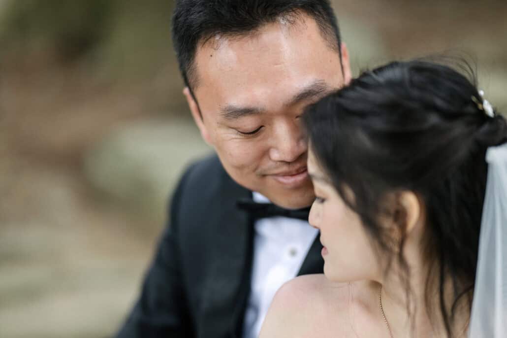 A close portrait of the groom smiling, his cheek against the bride's hair, both looking down, her pearl hair pin and veil at the edge.