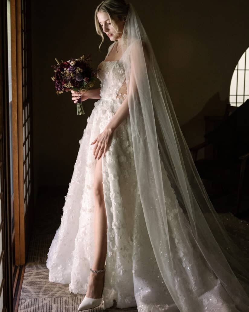 The bride stands by a shoji window in her floral wedding dress and long veil, holding a burgundy and purple bouquet, at a ryokan wedding in Japan.