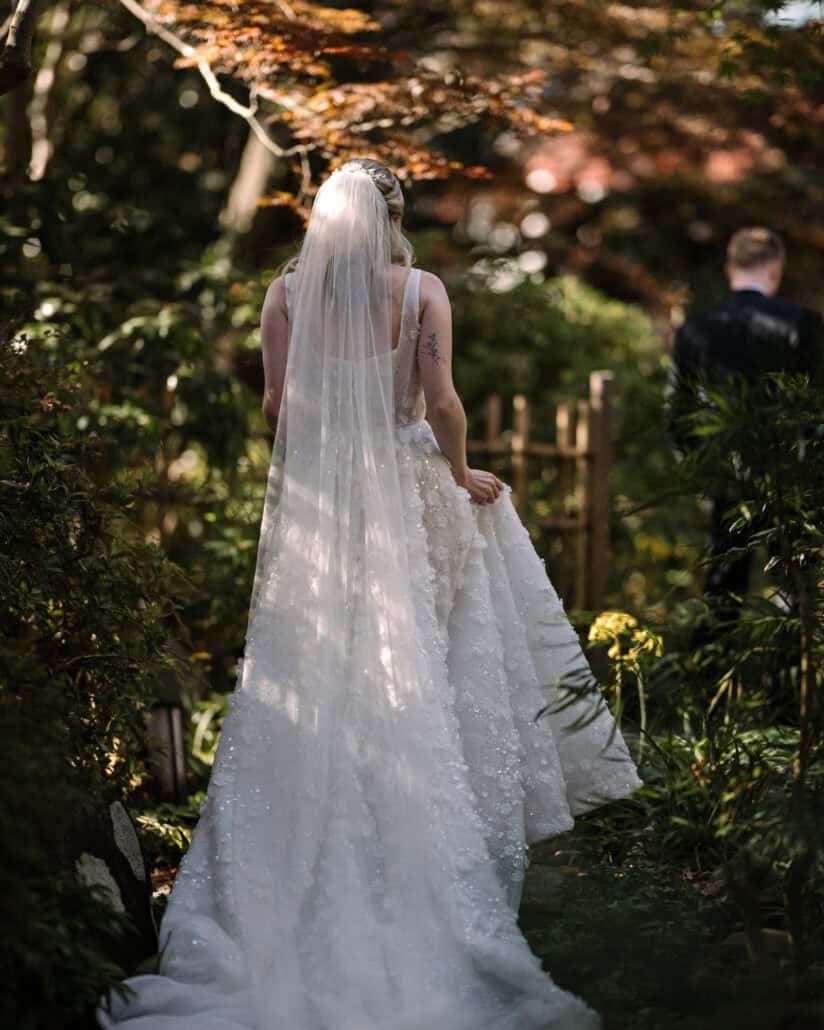 Komorebi through the maples dapples her veil as the bride walks to the groom, his back still turned, for a first look in a Japanese garden.