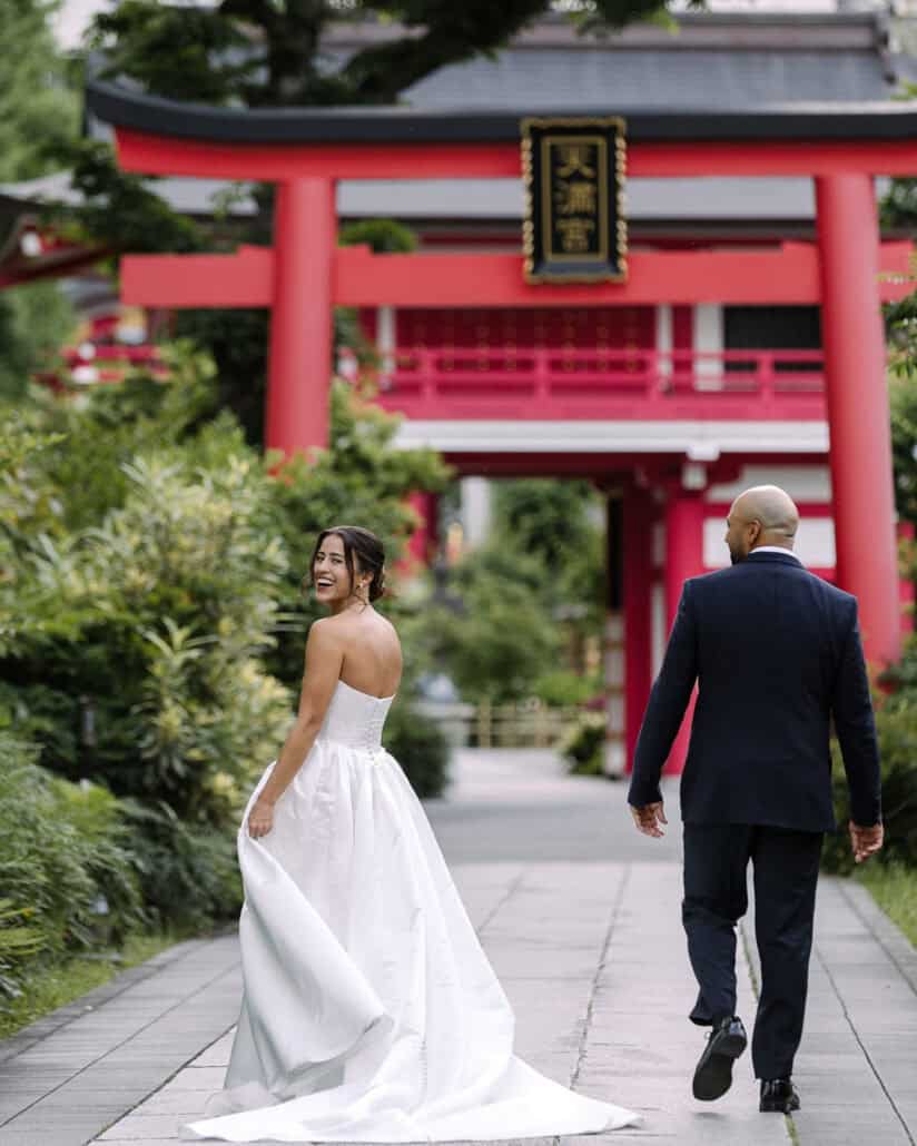 The bride glances back as she and the groom walk the stone path toward a vermilion torii at a Tokyo shrine.