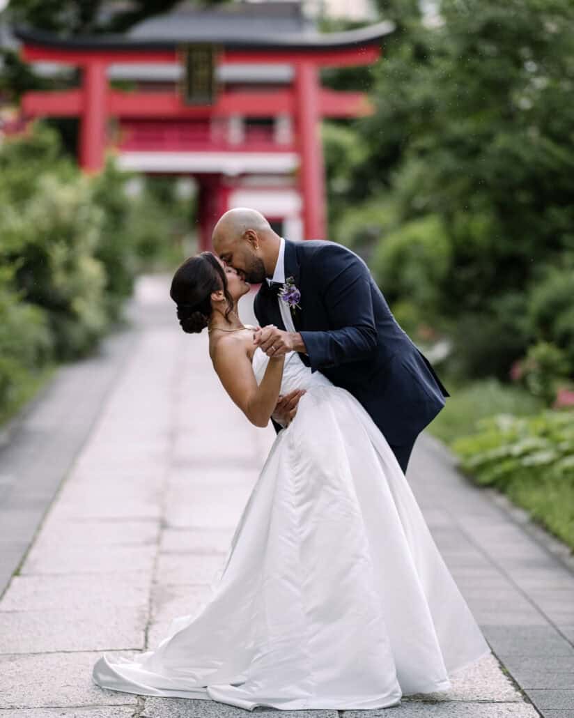 The groom dips the bride for a kiss on the stone path of a Tokyo shrine, a vermilion torii behind them.