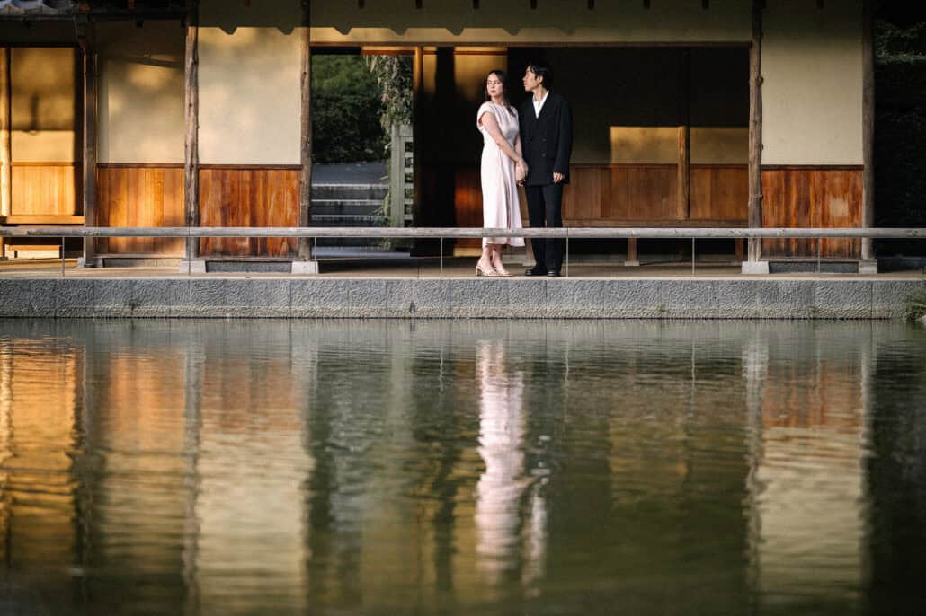 The couple stand on the veranda of a wooden pavilion above a still pond in Tokyo, the whole scene doubled in the water in golden light.