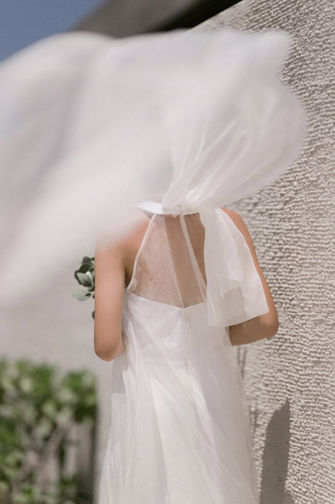 Seen from behind at a textured white wall, the bride's veil billows over her head above the bow of her halter dress, at her Miyakojima wedding.