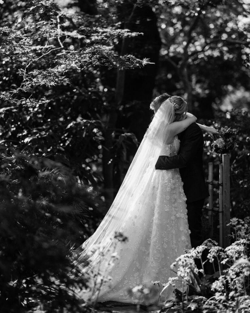 In black and white, the bride and groom hold each other by a bamboo fence, her veil trailing to the path, at their Japanese garden elopement.
