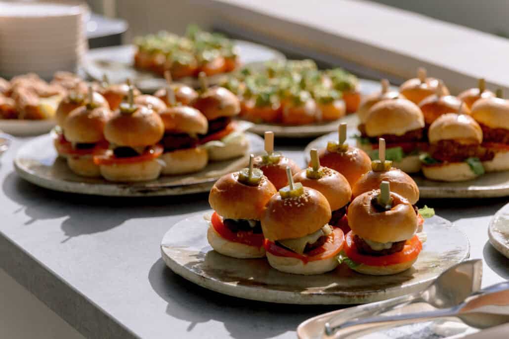 Mini burgers pinned with pickles on wooden picks wait on stoneware plates beside other small bites, for the wedding guests at Rosewood Miyakojima.