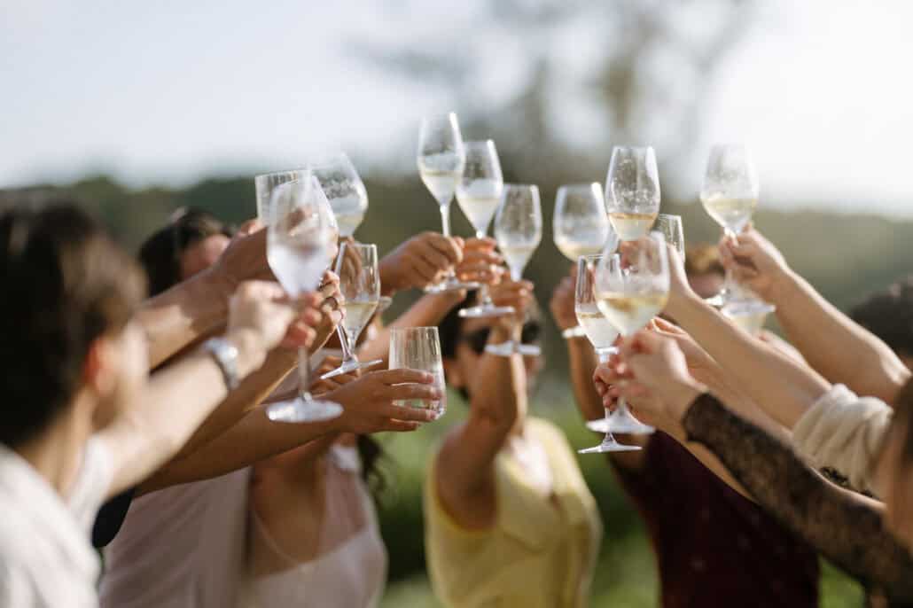 A ring of guests raise champagne glasses high against trees and pale sky, a yellow dress and a lace sleeve among them, at an Okinawa wedding party.