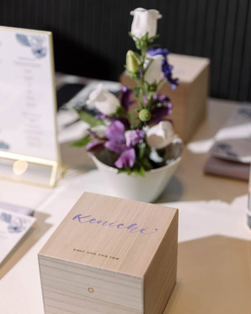 A small wooden gift box among purple flowers beside the menu, on a candlelit reception table at a Tokyo wedding.