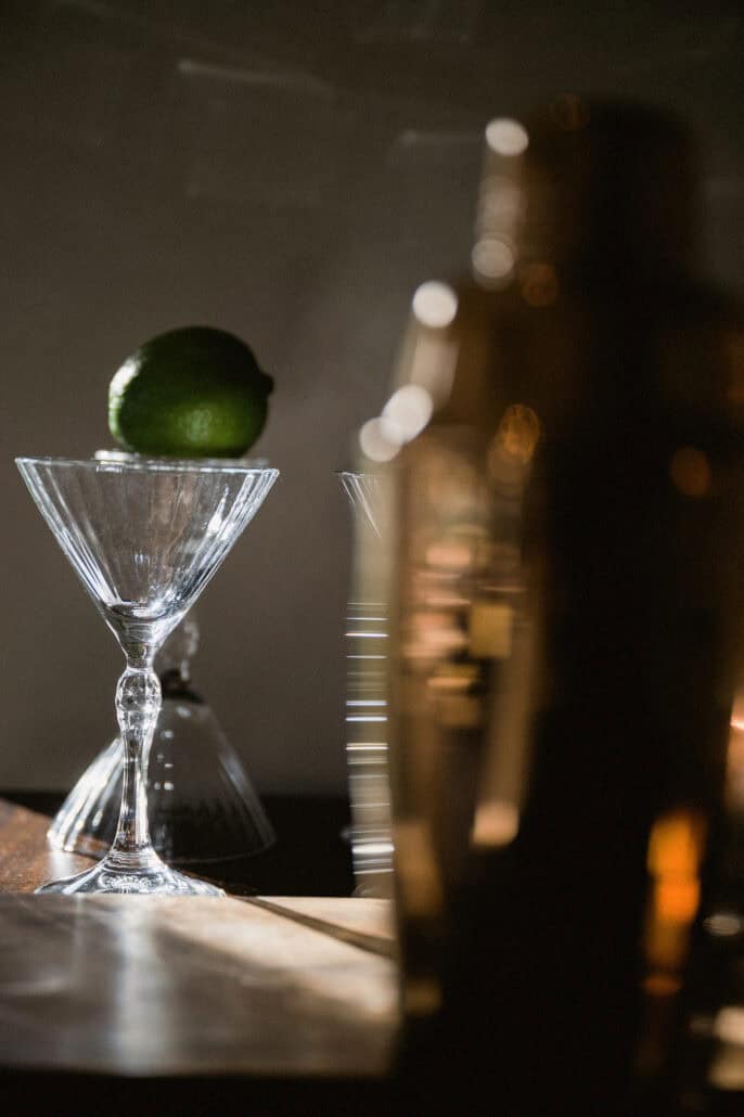 A lime balances on a cut-glass cocktail glass on a dark bar counter, bottles out of focus behind it, on a wedding evening at Rosewood Miyakojima.