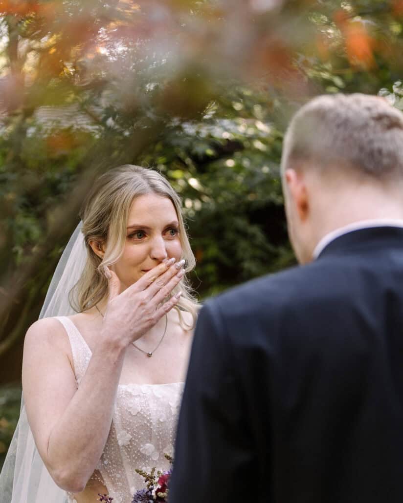 The bride covers her mouth with her hand, eyes wet, as she faces the groom at their Japanese garden wedding ceremony, framed by autumn maples.