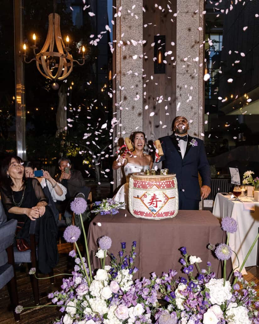 Petals fall over the couple as they open the sake barrel for the kagami biraki, guests filming, at their Tokyo wedding reception.