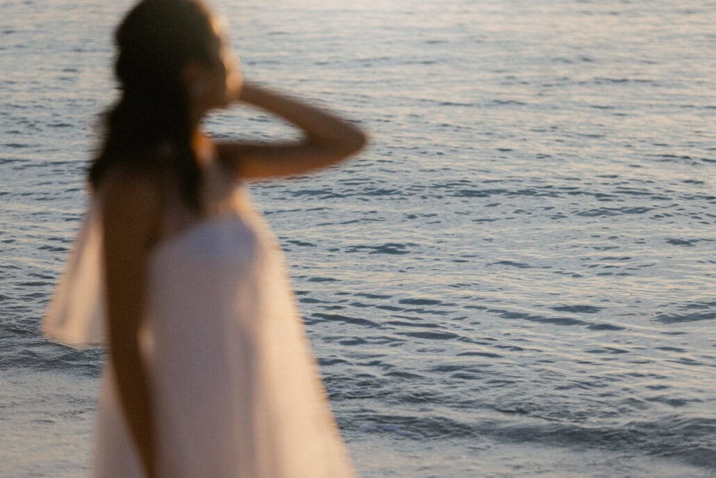 The bride stands soft in the foreground with a hand at her hair and sheer fabric at her shoulder, the sea glittering behind her in Miyakojima.