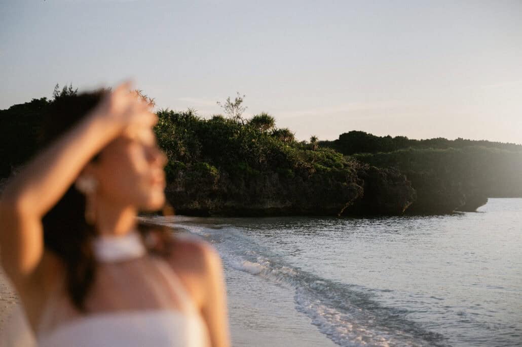 The bride shades her eyes, soft in the foreground, as small waves run up a white beach toward a wooded limestone headland on Miyako Island.