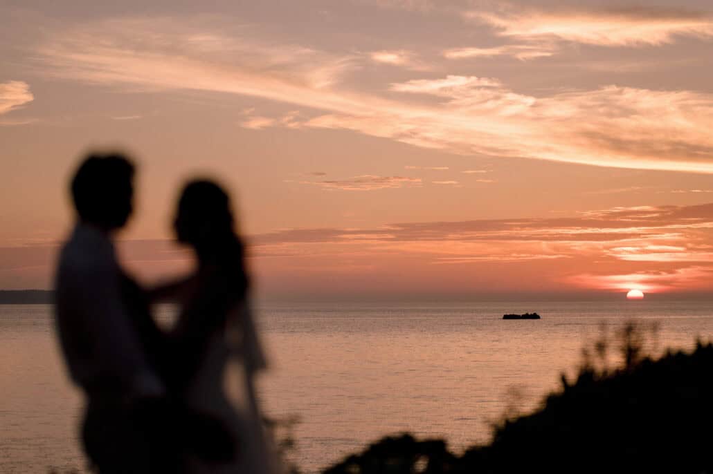 The couple hold each other as soft silhouettes while the sun meets the sea beside a low rock, pink cloud above, at their Miyakojima wedding.