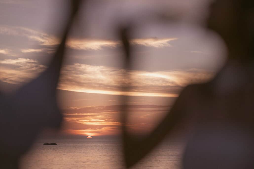 Blurred dark shapes in the foreground frame the sun as it touches the sea beside a low rock, streaks of cloud above, on a wedding evening in Okinawa.
