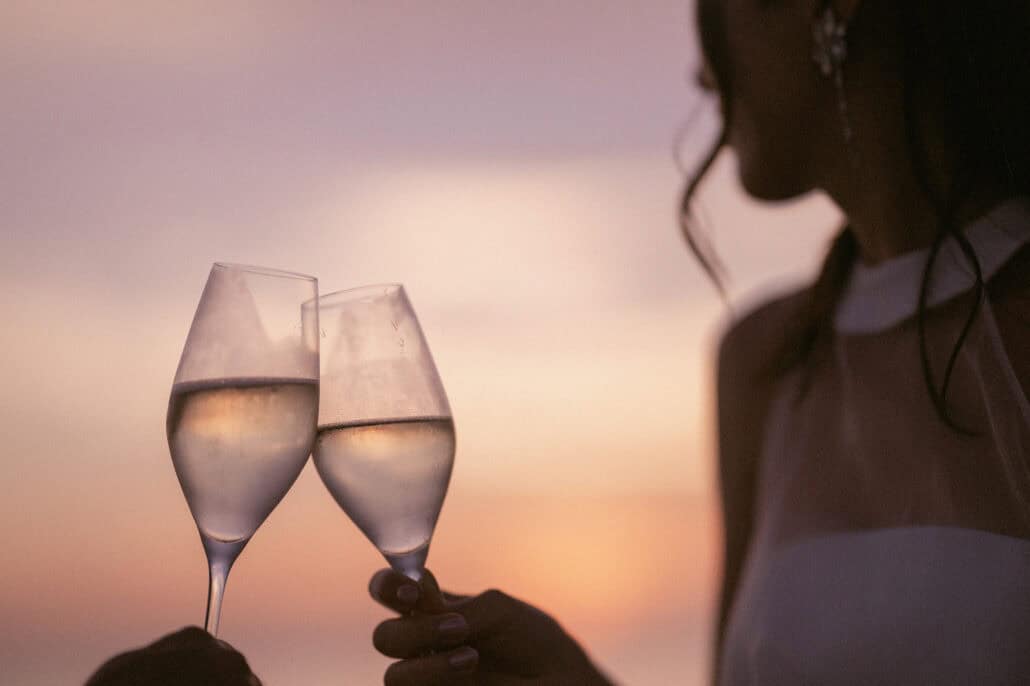 Two champagne glasses touch against a pink evening sky, the bride's shoulder and earring soft at the frame's edge, at an Okinawa destination wedding.