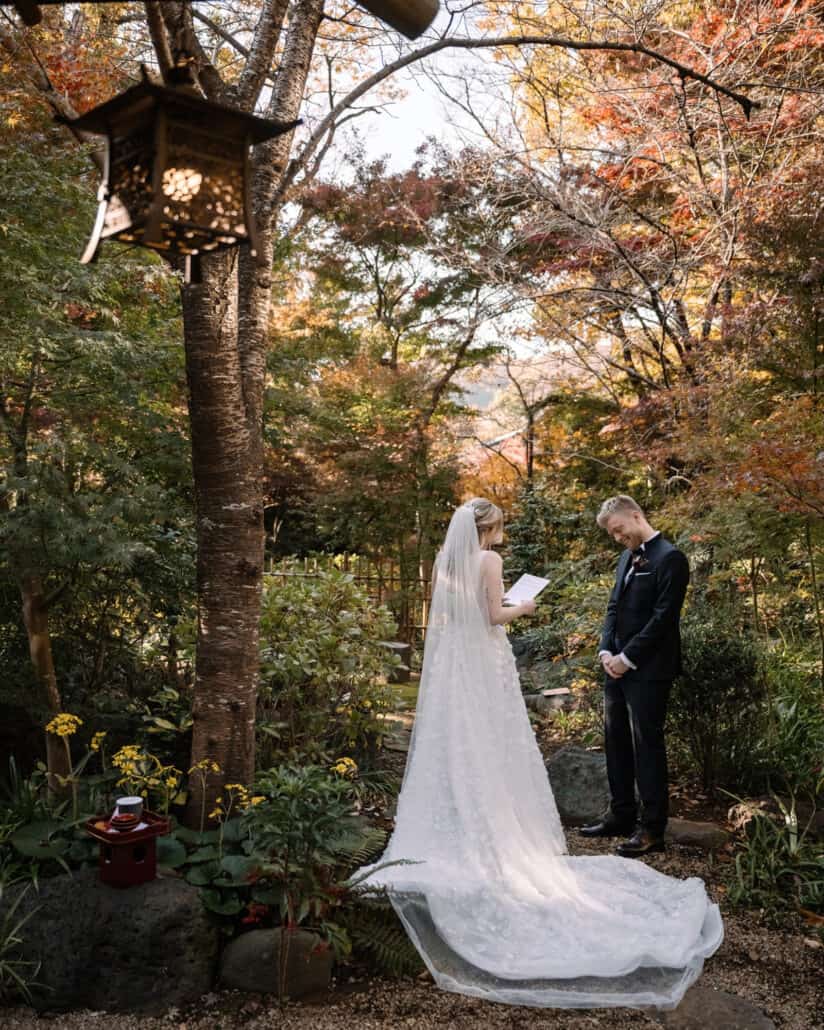 The bride reads her vows to the groom under autumn maples, a red sake set ready for the san san kudo, at a Japanese garden wedding ceremony.